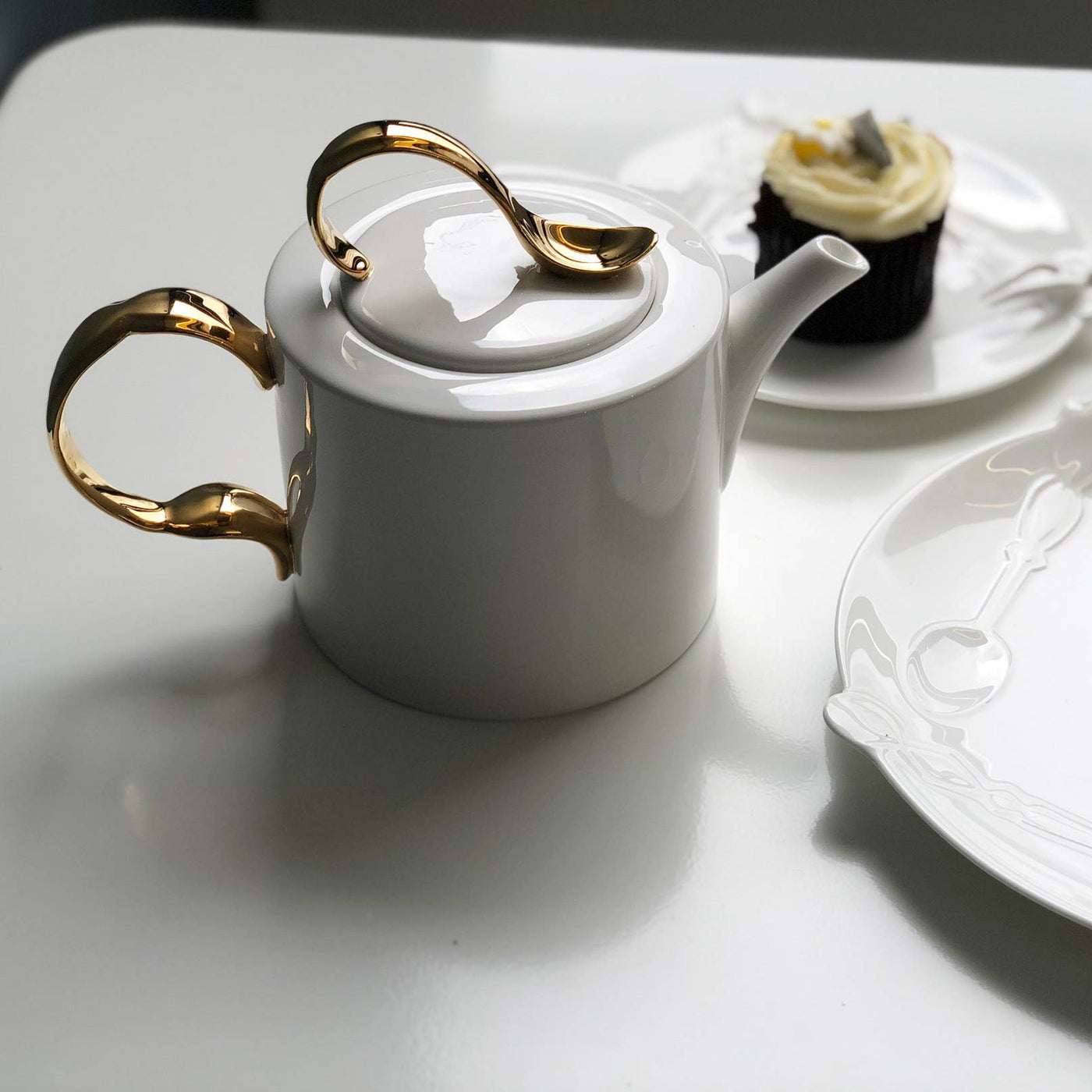White teapot with gold cutlery accents on a white surface with a cupcake and cutlery.